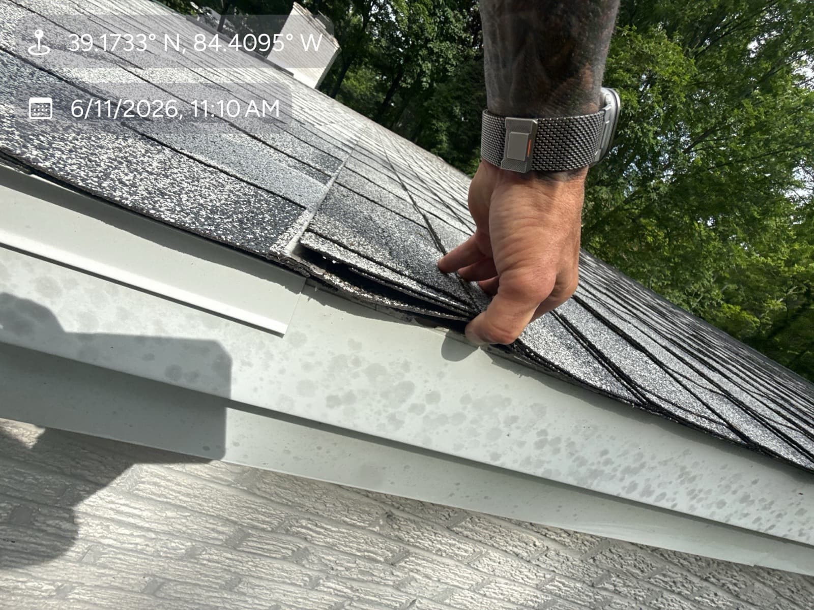 Inspector's hand lifting a hail-damaged shingle at the roof edge above a hail-spattered gutter